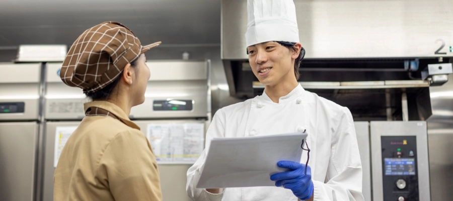 H.M.さん仕事風景　厨房の業務用の大きな冷蔵庫の前でスタッフと書類を見ながら会話している写真