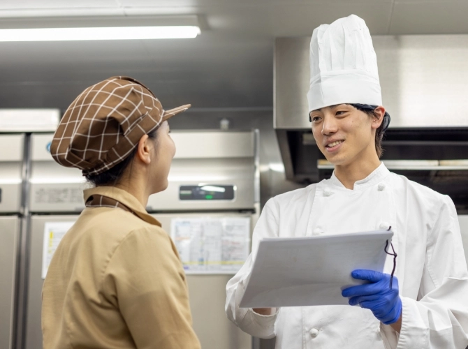 H.M.さん仕事風景　厨房の業務用の大きな冷蔵庫の前でスタッフと書類を見ながら会話している写真