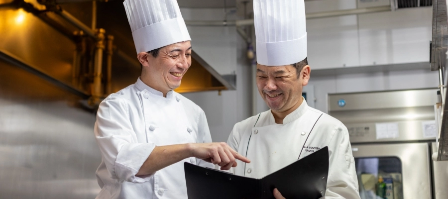 N.T.さん仕事風景　厨房で調理スタッフとメニューを見ながら笑顔で会話している写真