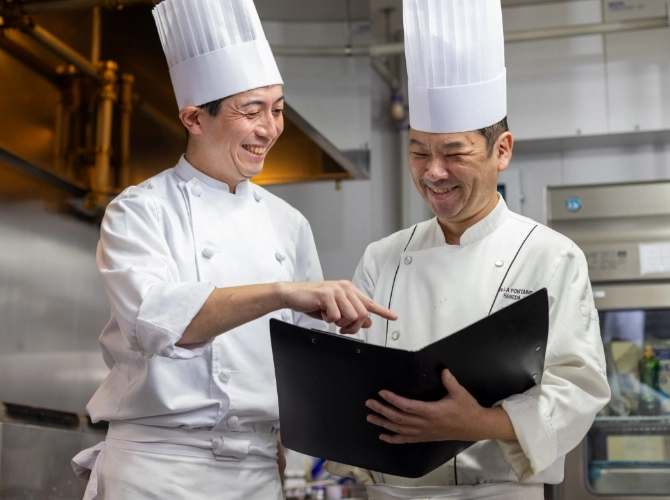N.T.さん仕事風景　厨房で調理スタッフとメニューを見ながら笑顔で会話している写真