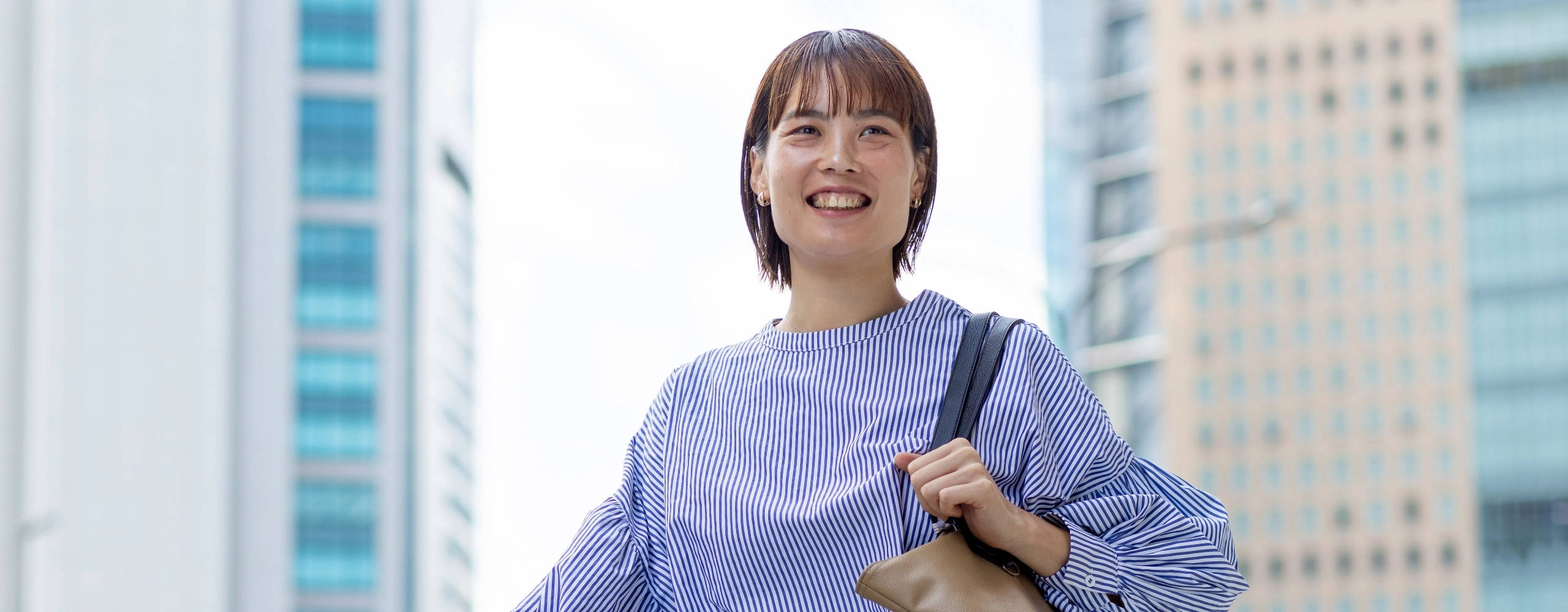 R.S.さんインタビューキービジュアル　高層ビルをバックにカバンを肩にかけて立っている写真