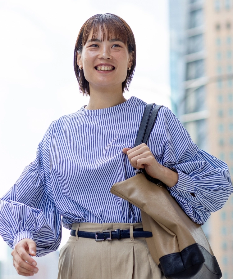 R.S.さんインタビューキービジュアル　高層ビルをバックにカバンを肩にかけて立っている写真